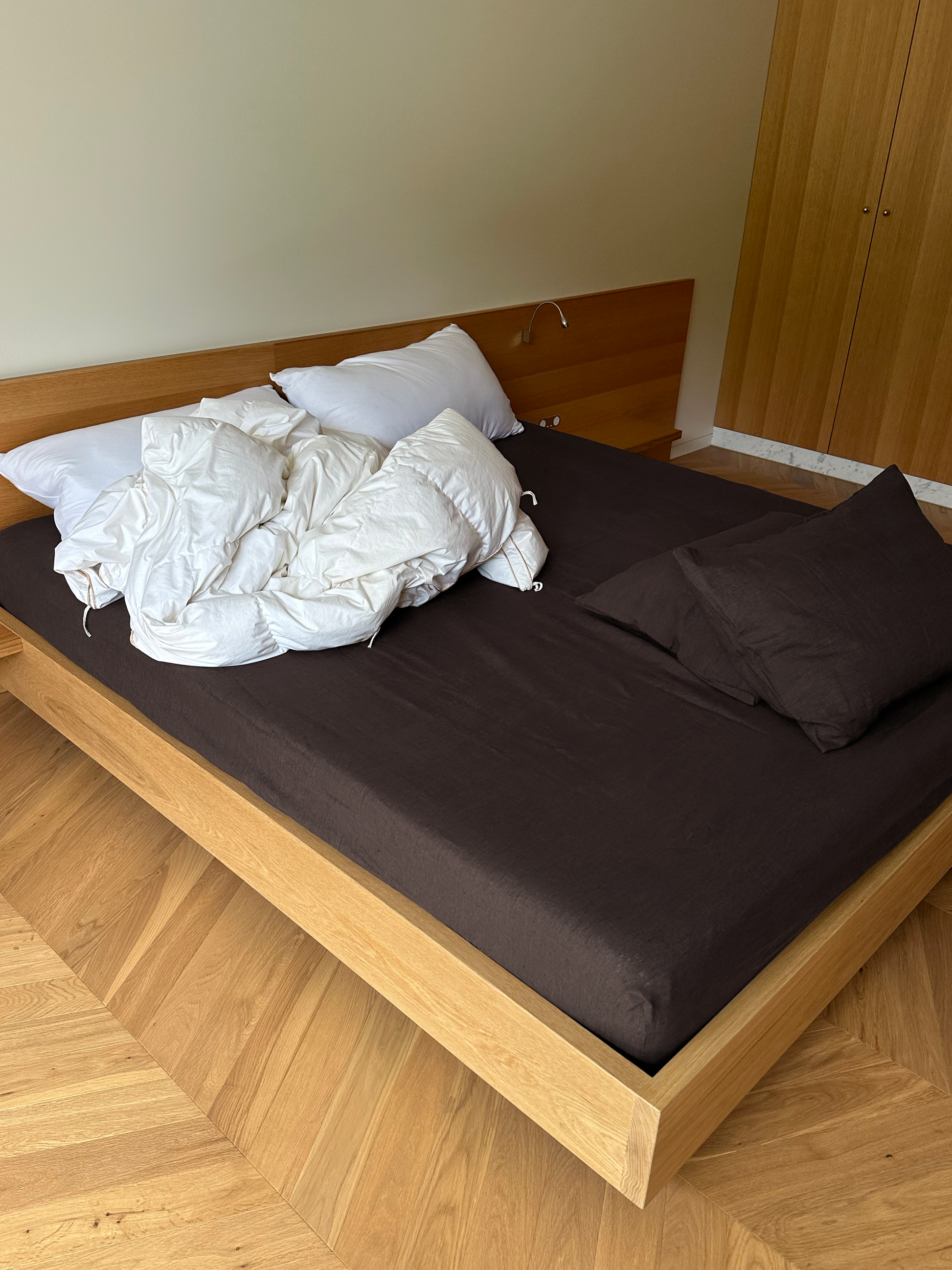 A modern wooden bed with Bark sheets, two dark pillows, and partially rumpled white bedding on the left. The room has wooden floors and a wooden wardrobe in the background, creating an eco-friendly atmosphere.