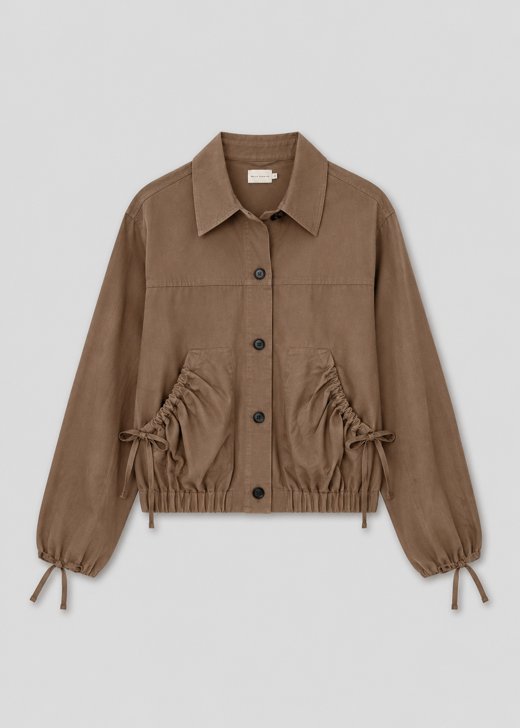 The Button Up Jacket - Brown is a light brown cropped jacket with a collar, front buttons, drawstring waist and cuffs, elastic hem, and two large front pockets, shown on a plain light background.