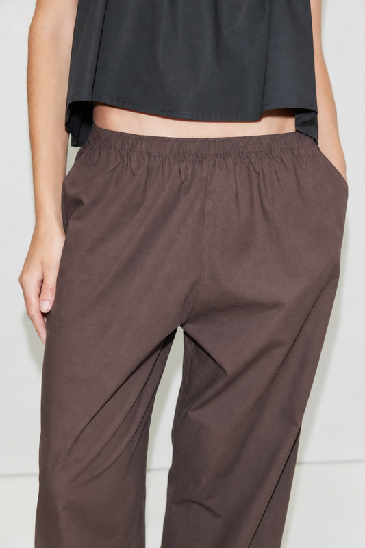 A person wearing the Ease Trouser - Toast by Deiji Studios, ankle length with a loose fit and elastic waistband, paired with a black top, shown from the torso down with one hand in their pocket.