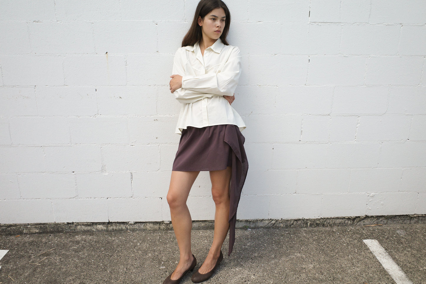 A woman stands against a white brick wall, arms crossed, wearing the Deiji Studios Cami Overshirt in Citrus Stripe, a short asymmetrical brown skirt with draped detail, and brown shoes. She gazes to the side with a neutral expression.