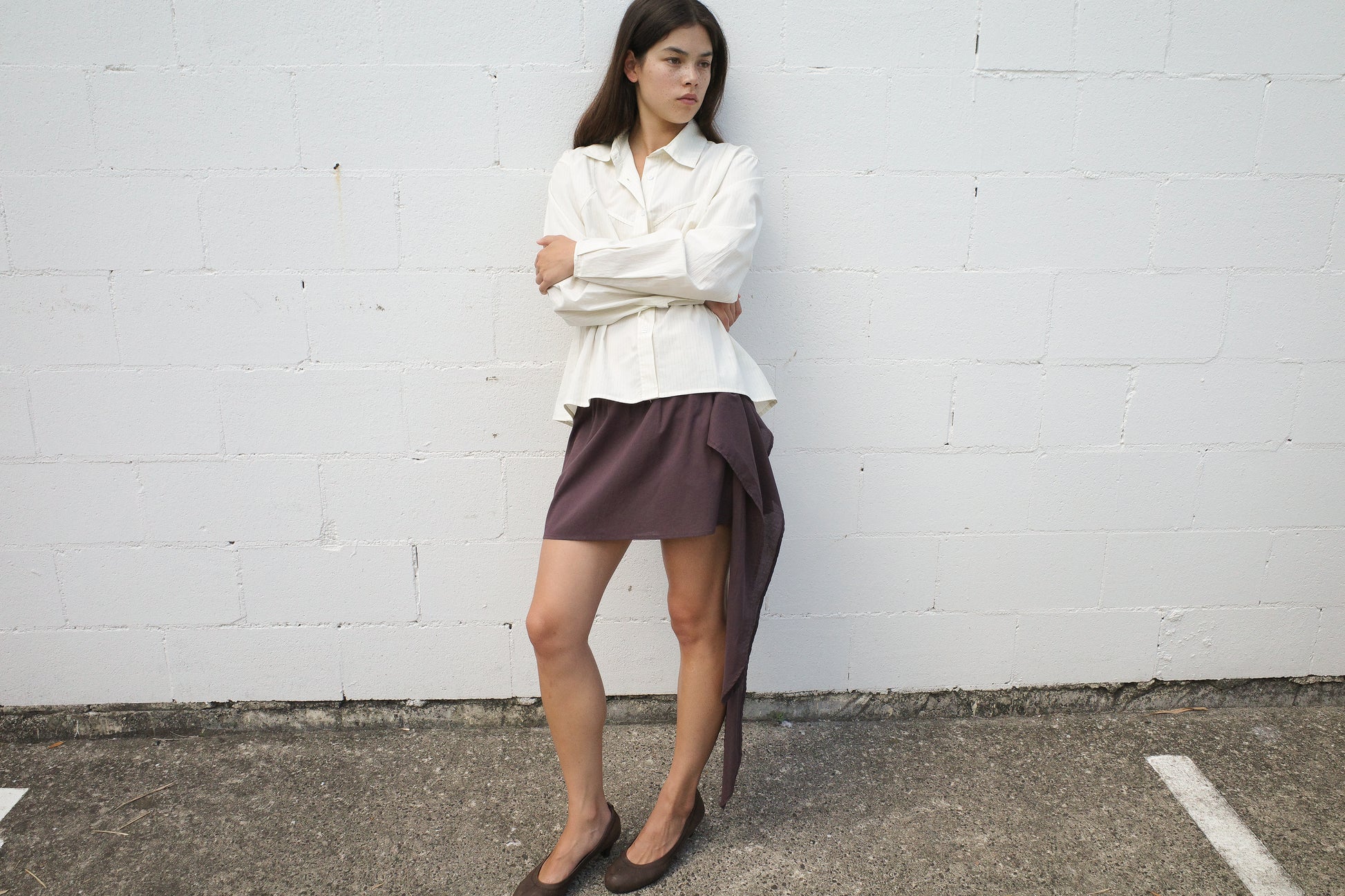 A woman stands against a white brick wall, arms crossed, wearing the Deiji Studios Cami Overshirt in Citrus Stripe, a short asymmetrical brown skirt with draped detail, and brown shoes. She gazes to the side with a neutral expression.