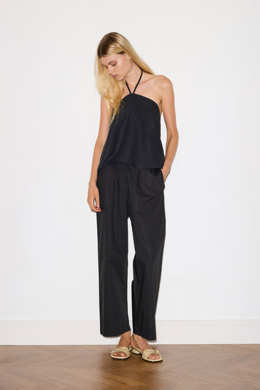 blonde haired female model wears a black relaxed halter top features flattering straight neckline, fine tie halter straps and snap closure at back with button detail, paired with a black ankle length pant features a soft minimal elastic waist with side seam pockets in a straight leg.