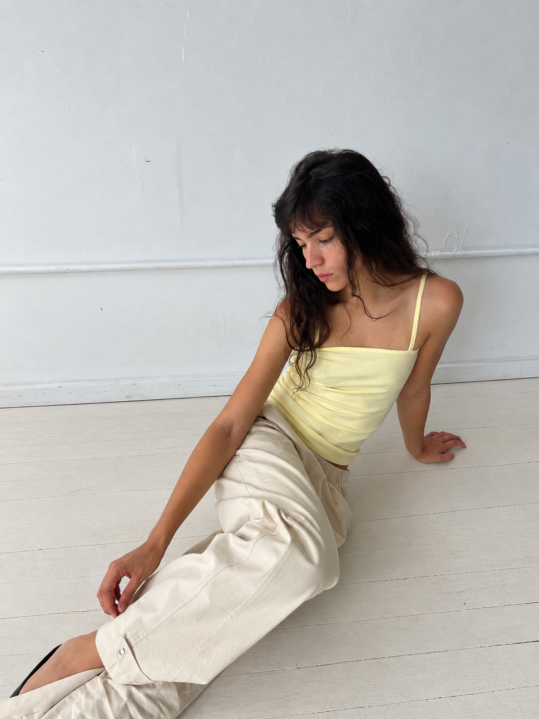 A woman sits on a white studio floor, wearing the Snap Tab Pant - Stone by Deiji Studios, a relaxed wide-leg pant with snap tabs at the hem, styled with the Scallop Tank in Lemon.