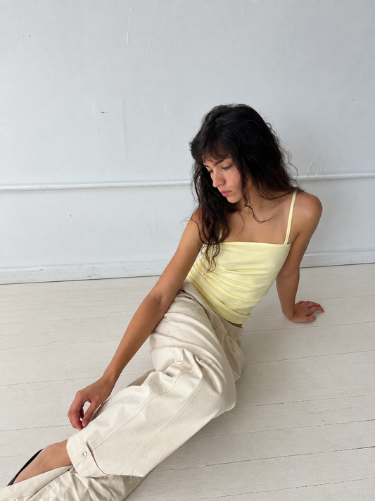 A woman sits on a white studio floor, wearing the Snap Tab Pant - Stone by Deiji Studios, a relaxed wide-leg pant with snap tabs at the hem, styled with the Scallop Tank in Lemon.