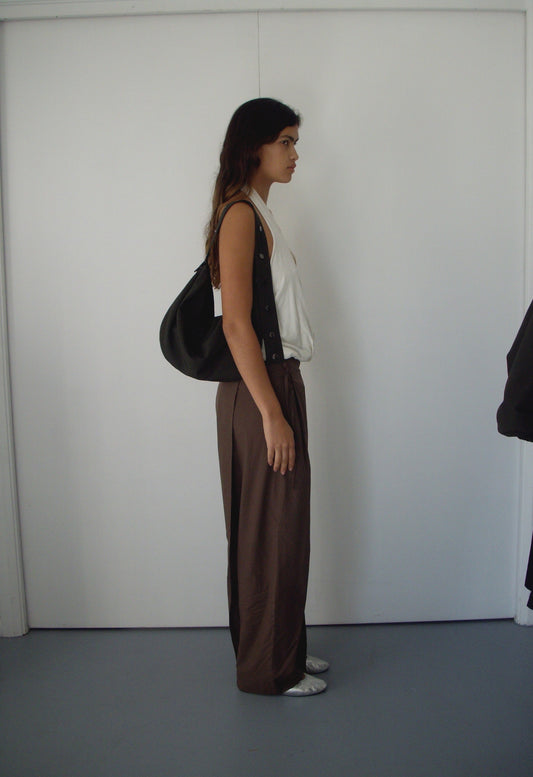 A person with long hair stands in profile against a white wall, wearing a white sleeveless top, loose brown pants from Deiji Studios, white shoes, and carrying the Press Stud Saddle Bag in Dark Chocolate.