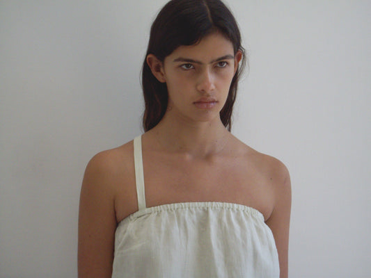 A person with long dark hair wears the Button Strap Top in Pastel Pistachio, made from organic French linen, standing against a white wall and looking slightly upward with a neutral or serious expression.