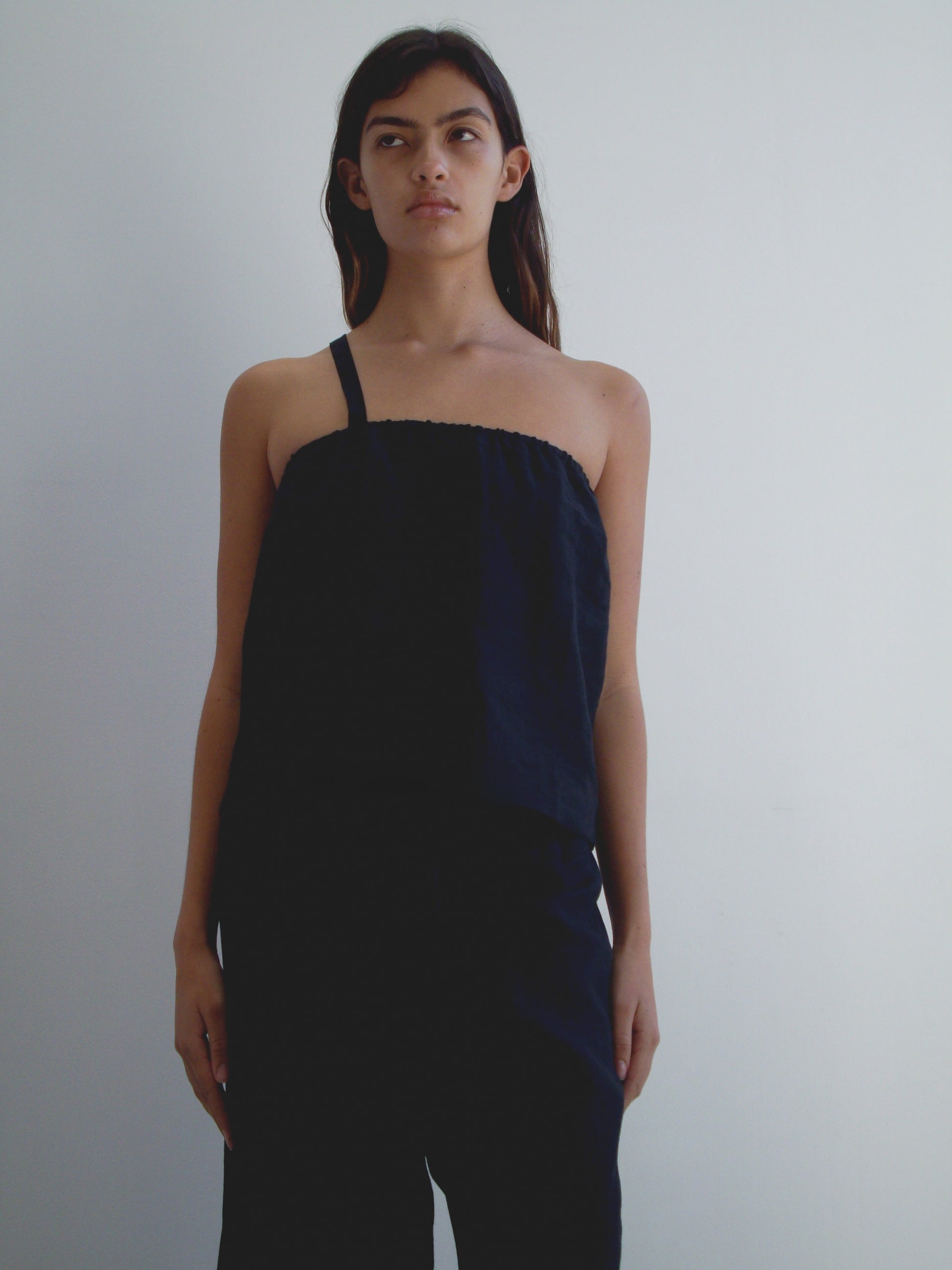 A woman with long dark hair stands against a white background, wearing the Button Strap Top - Black by Deiji Studios and black pants. She has a neutral expression and her arms rest at her sides.