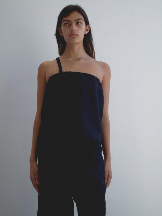 A woman with long dark hair stands against a white background, wearing the Button Strap Top - Black by Deiji Studios and black pants. She has a neutral expression and her arms rest at her sides.