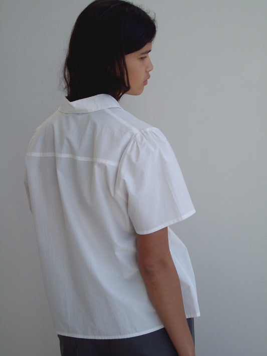 A person with shoulder-length dark hair stands sideways, facing away from the camera, wearing the Deiji Studios Sails Top in white and gray pants against a plain light gray background.