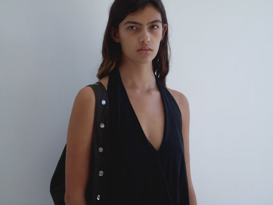 A woman with long dark hair wears the Jersey Top - Black from Deiji Studios, standing against a plain white wall with a neutral expression. She carries a bag with silver buttons over one shoulder.