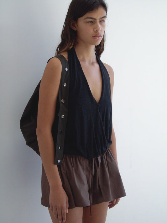 A person with long dark hair wears a sleeveless black halter top with side button details from Deiji Studios, loose brown shorts, and completes the look with the Press Stud Saddle Bag in Dark Chocolate, standing against a plain white wall.