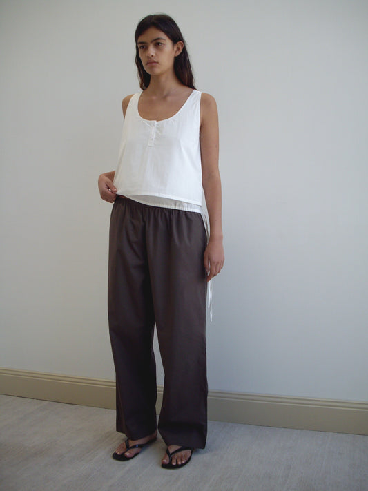 A woman with straight dark hair and a neutral expression stands indoors against a plain wall, wearing Deiji Studios Layered Top - White, wide-legged dark brown pants, and black sandals.