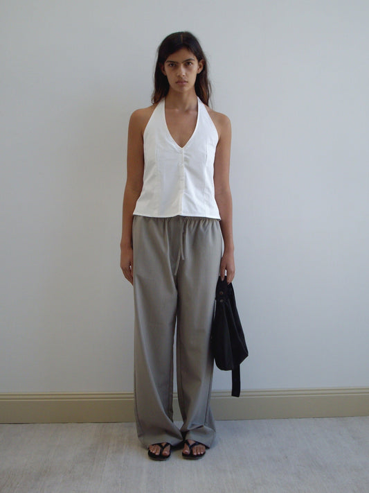 A woman stands against a plain wall wearing the Waist Halter - White, paired with loose gray trousers and black sandals. She holds a black item in her left hand and has a neutral expression.