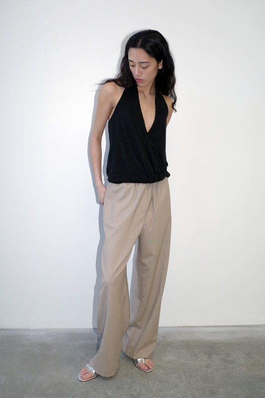 A woman with long dark hair stands against a white wall, looking down with her hands in her pockets. She wears a sleeveless black halter top, silver sandals, and Low Pocket Pure Wool Pant - Dove Stripe in a relaxed fit.