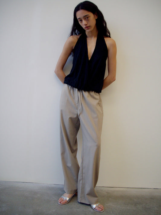 A person with long dark hair stands against a plain white wall, wearing a sleeveless black halter top, Low Pocket Pure Wool Pant - Dove Stripe, and silver open-toe sandals, with their hands behind their back.