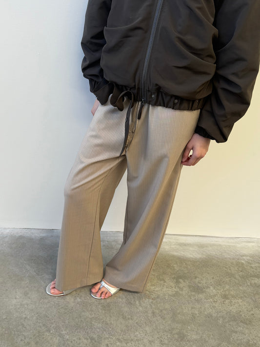 A person wearing Low Pocket Pure Wool Pant - Dove Stripe, a brown zip-up jacket, and white sandals stands on a concrete floor by a light-colored wall. Their face is not visible.