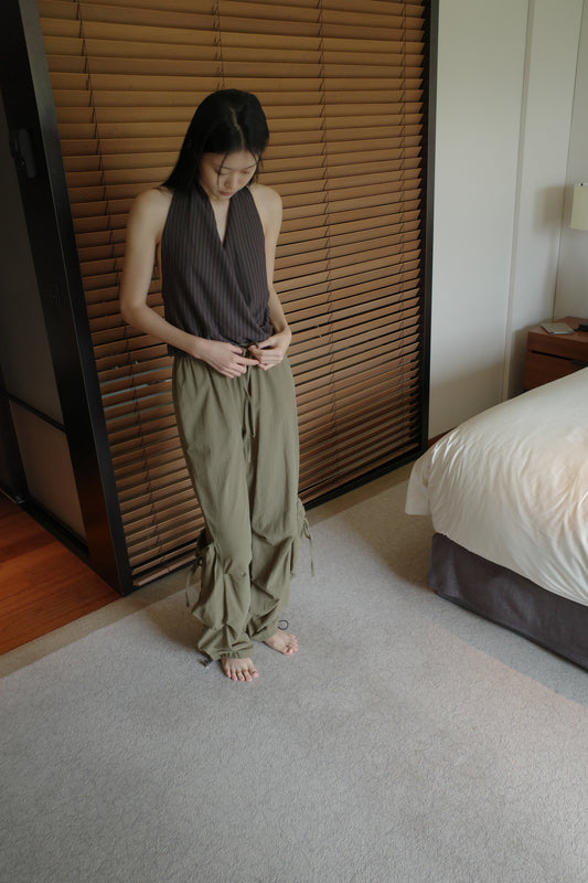 A woman with long dark hair stands barefoot in front of wooden blinds in a bedroom, wearing the Drawcord Pocket Pant - Khaki by Deiji Studios, khaki wide-leg trousers, with a brown striped halter top.