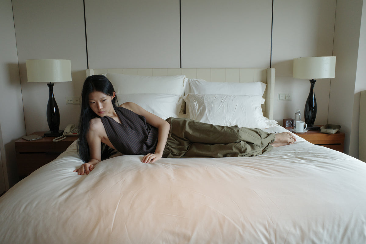 A woman with long dark hair reclines on a large white bed in a softly lit room, wearing the Drawcord Pocket Pant - Khaki by Deiji Studios, khaki wide-leg trousers, with a brown striped halter top.