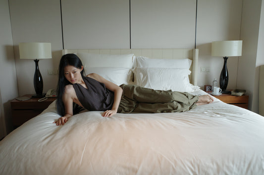 A woman with long dark hair reclines on a large white bed in a softly lit room, wearing the Drawcord Pocket Pant - Khaki by Deiji Studios, khaki wide-leg trousers, with a brown striped halter top.