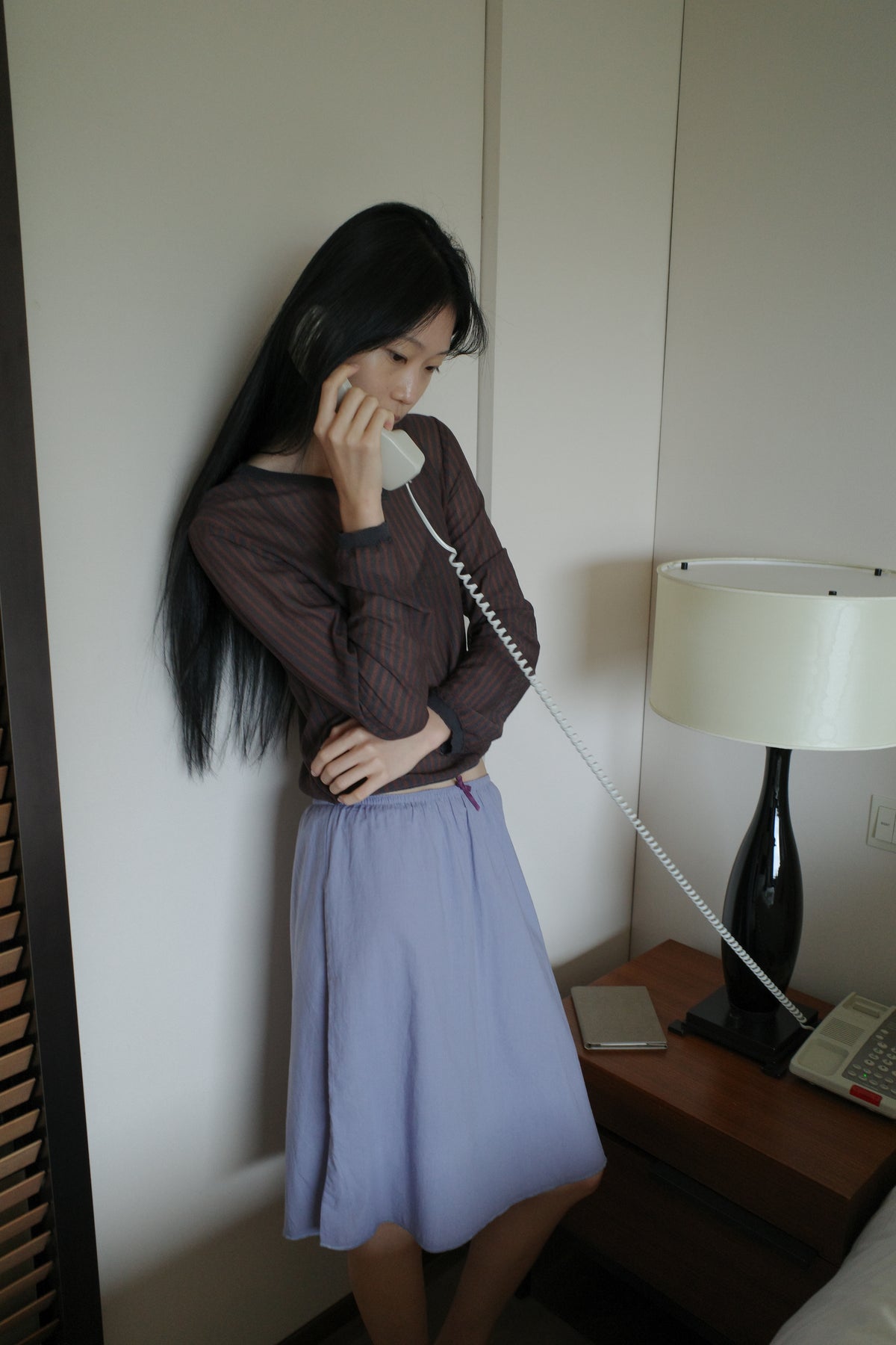 A woman with long dark hair leans against a wall in a softly lit room, holding a vintage telephone handset to her ear and looking down, wearing a sheer brown striped long-sleeve top and the Basic Skirt - Thistle by Deiji Studios, a periwinkle cotton voile midi skirt.