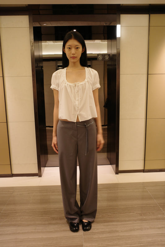 A woman with long dark hair stands in front of a lift wearing the Flounced Top - White by Deiji Studios, a white top with a frilled scoop neckline, front tie and puff sleeves, with grey pinstripe trousers and black shoes.