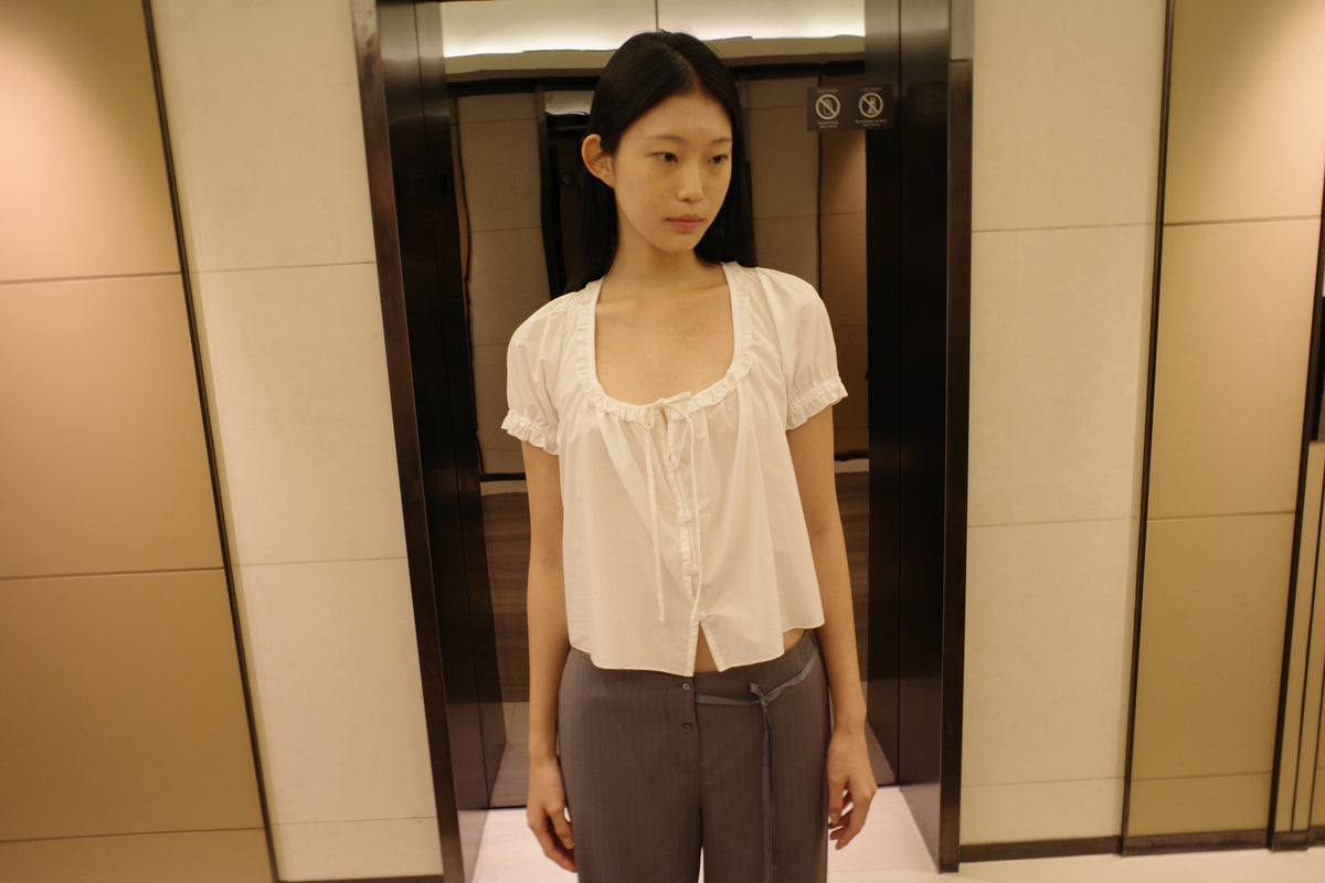 A woman with long dark hair stands in front of a lift wearing the Flounced Top - White by Deiji Studios, a white top with a frilled scoop neckline and puff sleeves, with grey pinstripe trousers.