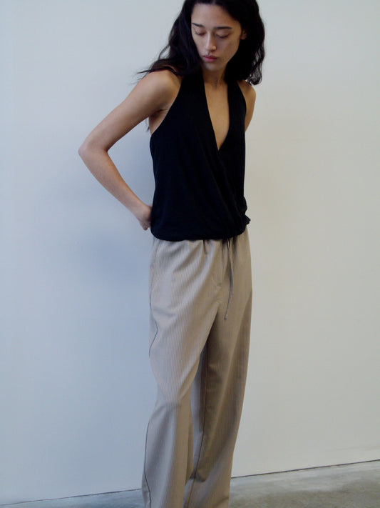 A person with long dark hair stands against a white wall, wearing a sleeveless black V-neck top and the Low Pocket Pure Wool Pant - Dove Stripe, looking down with hands behind their back.