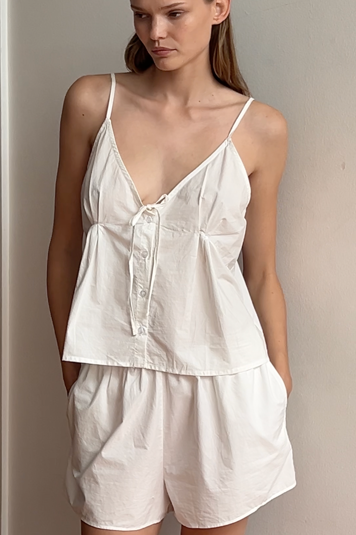 A woman stands indoors wearing the Placket Short Set - White with a loose, sleeveless fit in organic cotton. Her hands are in her pockets as she gazes slightly to the side.