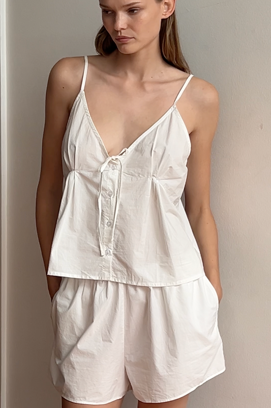A woman stands indoors wearing the Placket Short Set - White with a loose, sleeveless fit in organic cotton. Her hands are in her pockets as she gazes slightly to the side.