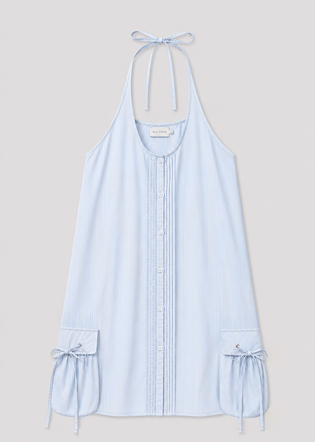 The Pocket Halter Dress - Mist Stripe by Deiji Studios is a sleeveless, light blue pinstriped halter dress with a button-down front and large shirt cuff details at the hem, each tied with a bow.