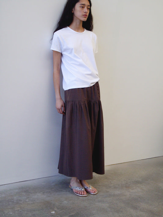 A person with long dark hair wears a white t-shirt, the Deiji Studios Button Skirt in Chocolate, and sandals while standing against a plain white wall on a concrete floor.
