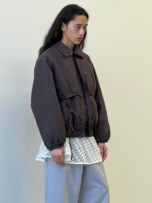 A person with long dark hair stands against a pale background, wearing the Double Layered Jacket - Slate Check over a white textured top and light gray pants, looking thoughtfully to the side.