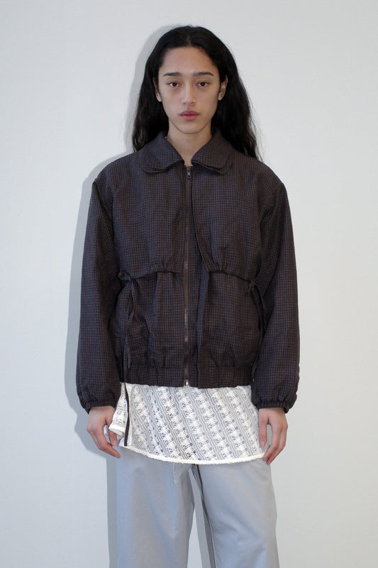 A person with long dark hair stands against a plain light wall, wearing the Double Layered Jacket - Slate Check over a white patterned shirt and light gray pants, looking directly at the camera with a neutral expression.