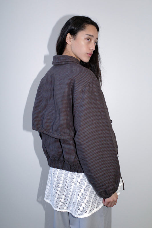 A person with long dark hair stands sideways, wearing the Double Layered Jacket - Slate Check over a white patterned shirt and gray pants, set against a plain light gray background.