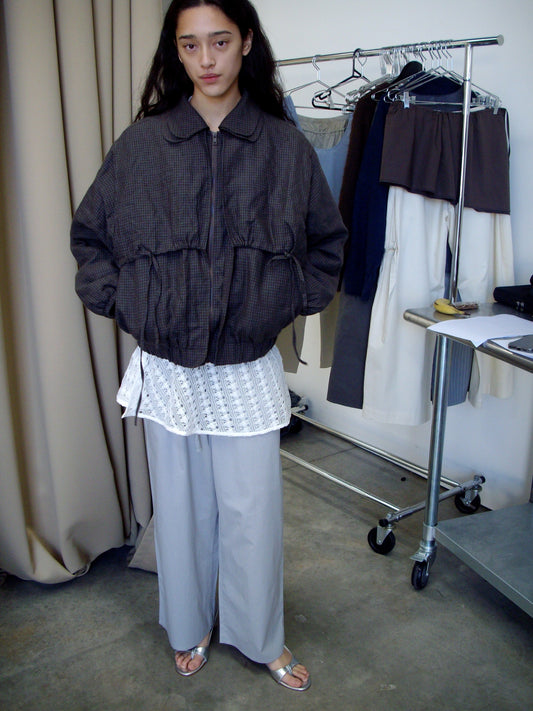 A person with long dark hair wears a loose Double Layered Jacket - Slate Check, a white lace top, wide light gray pants, and silver sandals. Behind them is a clothing rack with hanging garments and a curtain to the left.