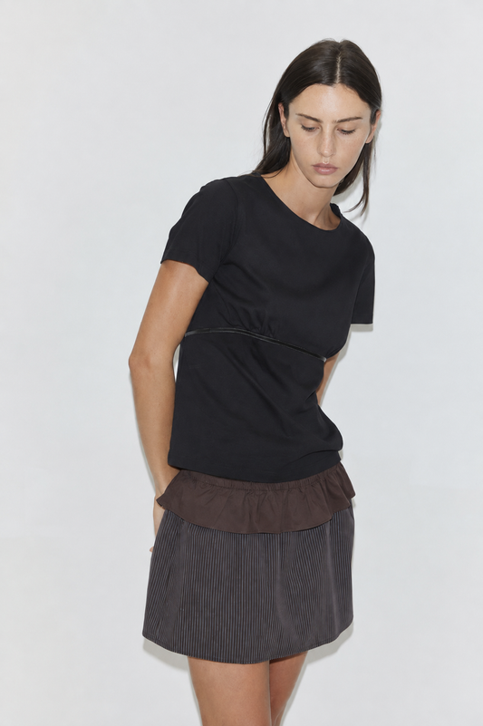 A woman with long dark hair stands against a plain white background looking down, wearing the Flounce Layer Mini Skirt - Brown Blue Stripe by Deiji Studios, a fine brown-blue stripe mini skirt with a brown flounce waist, and a black tee.