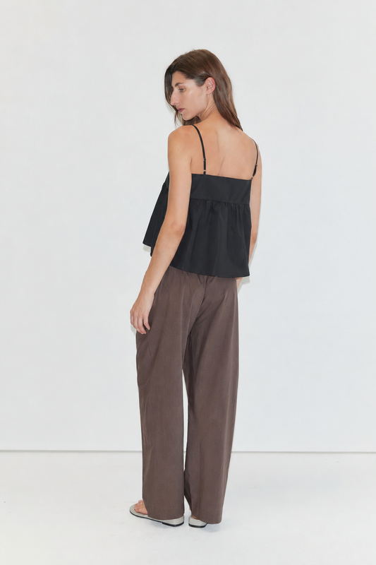 A woman with long brown hair stands against a plain white background, wearing a black sleeveless top with thin straps and the Ease Trouser - Toast from Deiji Studios.