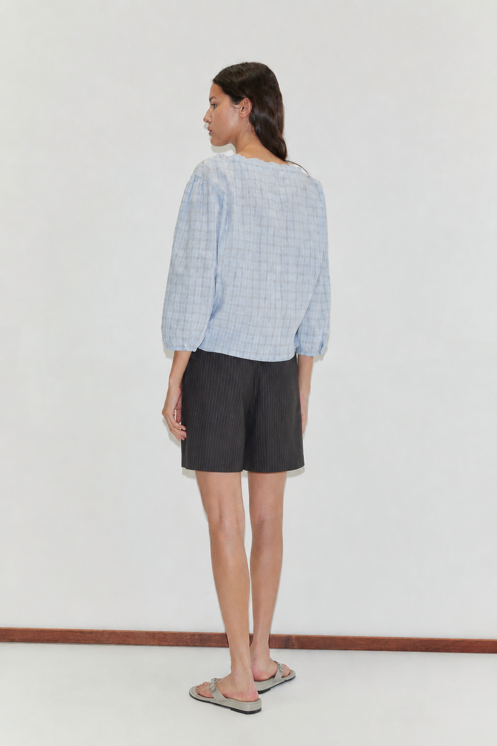 A woman with long hair, facing away, wears the Corded Top - Powder Check from Deiji Studios, dark knee-length shorts, and flip-flops against a plain white backdrop.