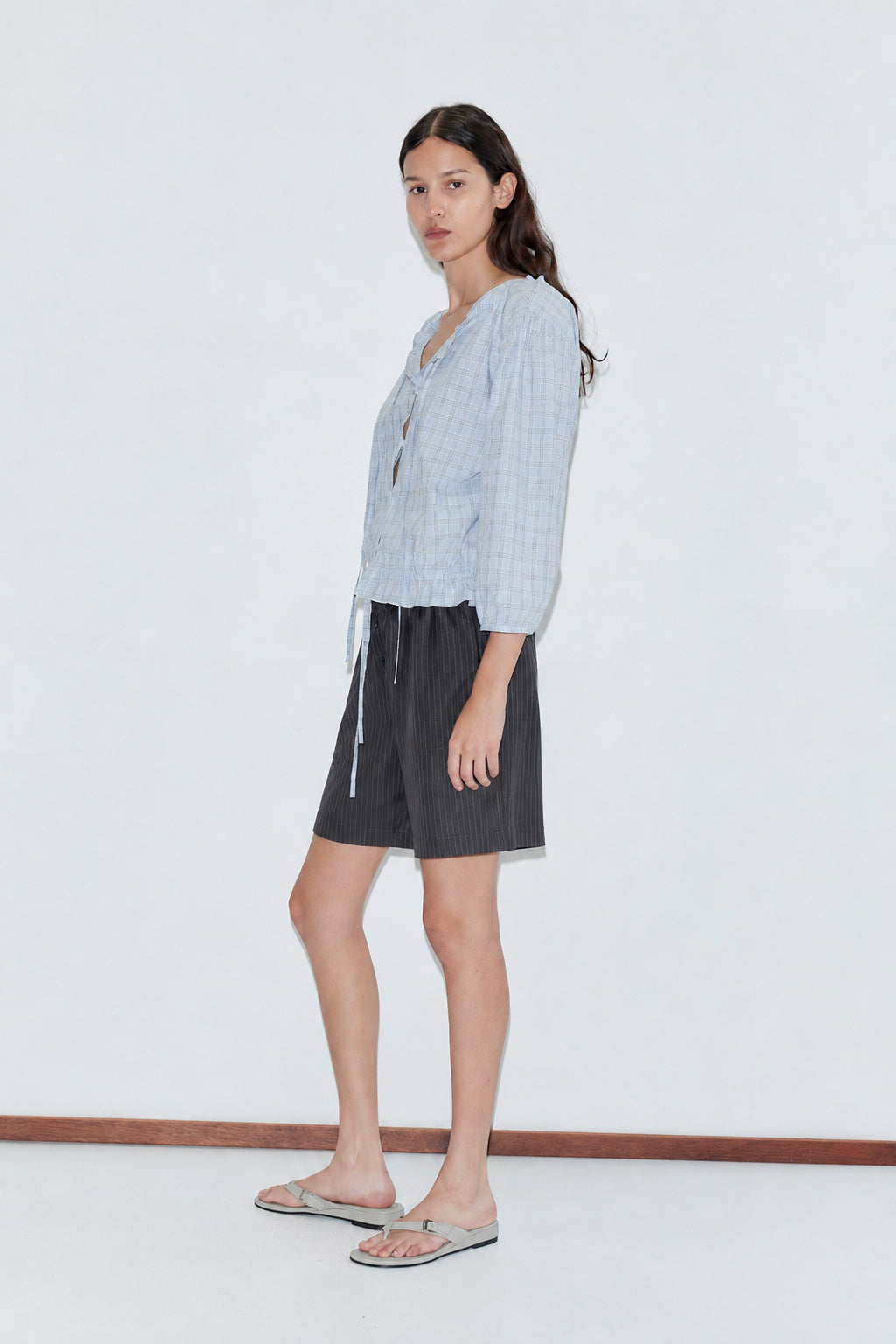 A woman with long dark hair stands sideways against a white background, wearing the Corded Top - Powder Check by Deiji Studios, dark gray knee-length shorts, and light-colored flip-flops.