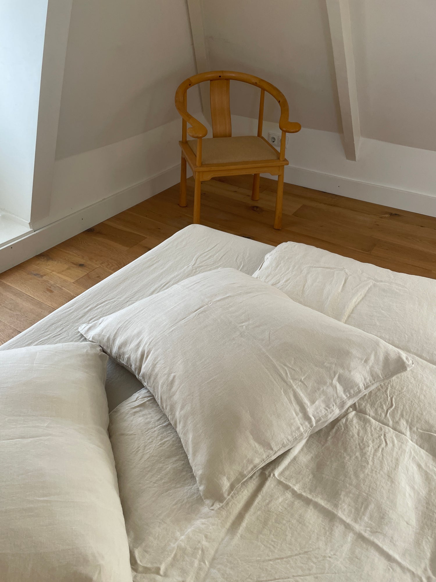 A neatly made bed with off-white linens and Pillow Slips, a pillow in the foreground, and a wooden chair in the corner of a minimalist room with wooden flooring.
